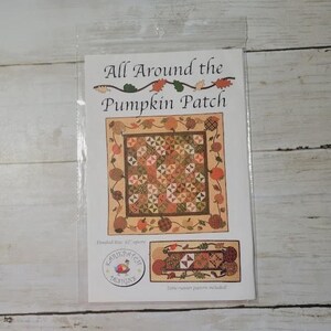 May include: A package featuring a quilt pattern titled "All Around the Pumpkin Patch." The design includes a central patchwork square surrounded by pumpkins and leaves. The finished size is 62 inches square. A table runner pattern is also included.
