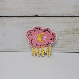 May include: A pink cloud-shaped patch with a yellow crescent moon and rain drops. The patch has a white border and black stitching details. The rain drops are yellow. This patch is suitable for clothing and accessories.