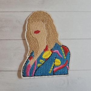 May include: Embroidered patch featuring a stylized portrait of a person with blonde hair and red lips. The person is wearing a blue jacket with yellow, pink, and red embroidered details. The patch has a white border and is likely for clothing or accessories.