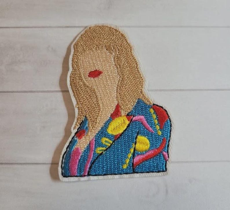 May include: Embroidered patch featuring a stylized portrait of a person with blonde hair and red lips. The person is wearing a blue jacket with yellow, pink, and red embroidered details. The patch has a white border and is likely for clothing or accessories.