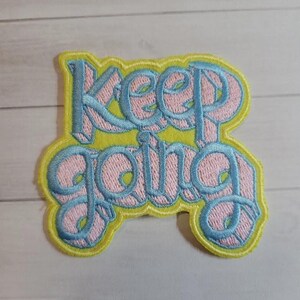 May include: Embroidered patch with the words "KEEP GOING" in light blue script over a pink background, outlined in yellow. The patch is rectangular with rounded edges, suitable for clothing or accessories.