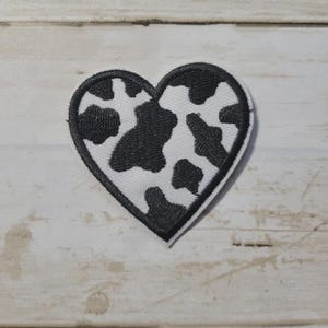 May include: Heart-shaped patch with a black outline and a white background featuring black cow spots. The patch is likely made of fabric and is designed to be sewn or ironed onto clothing or accessories.