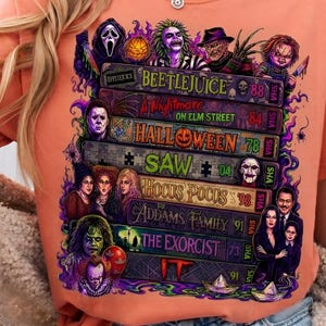 May include: A peach-colored t-shirt featuring a graphic print of stacked VHS tapes labeled with horror movie titles including Beetlejuice, A Nightmare on Elm Street, Halloween, Saw, Hocus Pocus, The Addams Family, The Exorcist, and It, with characters.