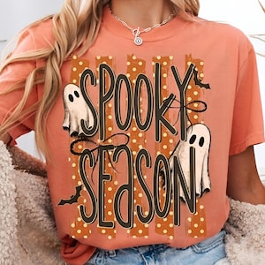Op de afbeelding: Een koraalkleurig t-shirt met korte mouwen en de tekst "SPOOKY SEASON" in donkerbruine letters. Het ontwerp bevat twee witte spookillustraties, kleine zwarte vleermuizen en verticale oranje strepen met witte stippen op de voorzijde.