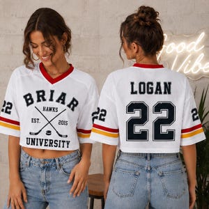 May include: White mesh crop top with red and yellow accents. The front features "Briar Hawks University" text and crossed hockey sticks. The back displays "Logan 22".