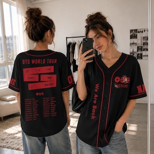 May include: Black baseball jersey with red trim and text. The jersey features "BTS WORLD TOUR" on the back with tour dates and "We Are Back" on the front. The jersey has a V-neck and short sleeves.