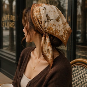 May include: A square silk headscarf featuring a cream background with a brown border and a pattern of flying ducks and autumn leaves in shades of brown and gold. The scarf is tied under the chin, displaying a detailed nature-inspired print.