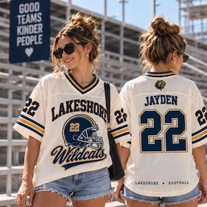 May include: An off-white V-neck football jersey featuring navy blue and gold accents. The front displays 'LAKESHORE WILDCATS' with a helmet graphic and the number 22. The back shows 'JAYDEN', the number 22, and the text 'LAKESHORE FOOTBALL'.