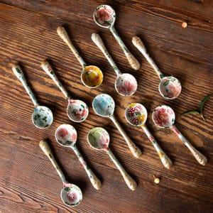 May include: A selection of ceramic spoons, each adorned with a hand-painted floral design. The spoons display a variety of floral patterns in pink, red, yellow, and blue hues. They rest on a wooden surface, with a cream-colored base.