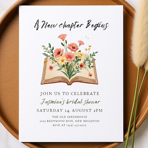 May include: A white invitation card with the text "A New chapter Begins" and details for Jasmine's bridal shower. The card features an open book illustration with floral arrangements. The card is on a wooden tray with decorative wheat stalks and a white ribbon.