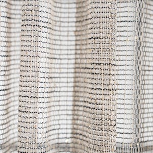 May include: A sheer curtain fabric featuring a vertical striped pattern created by alternating bands of black and beige open-weave mesh. The textile incorporates subtle metallic gold threads woven throughout the grid structure, adding a reflective quality to the light-coloured material.