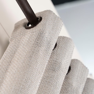 May include: A beige curtain with a textured, woven fabric finish hanging from a dark brown rod. The curtain features evenly spaced, circular dark brown grommets along the top edge, allowing the fabric to fold into consistent, vertical pleats.