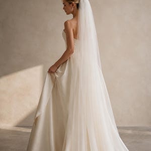 May include: A floor-length ivory wedding dress with a strapless bodice and a full, flowing skirt. The garment is paired with a long, sheer, single-tier tulle veil that extends down the back, creating a soft, layered silhouette with a clean finish.