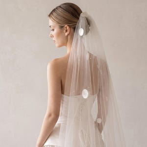 May include: A sheer white bridal veil featuring scattered circular white sequins and small pearl-like accents. The veil is attached to a hair comb and drapes down the back, showcasing a delicate tulle fabric with a soft, translucent appearance.