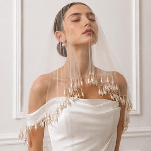 Puede incluir: Un velo de novia blanco y transparente con un borde festoneado adornado con numerosas perlas artificiales colgantes en forma de lágrima. El velo se coloca sobre la cabeza y los hombros, descansando sobre una prenda de satén blanco.
