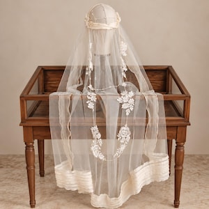 May include: A sheer ivory bridal veil featuring delicate white floral lace appliques and small pearl accents. The veil is draped over a mannequin head, showcasing a layered tulle design with a pleated fabric border along the bottom edge.