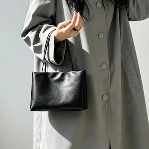 May include: A smooth black rectangular handbag with two thin top handles, held against a light gray long-sleeved coat featuring a button-down front, a classic collar, and cuffed sleeves, creating a clean and structured appearance.