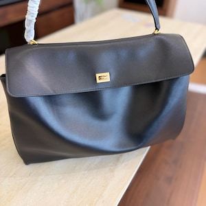 May include: Black leather handbag with a flap closure and a top handle. The bag features a gold-toned rectangular clasp on the front. The handle is attached with gold-toned hardware. The bag is sitting on a light-colored surface.