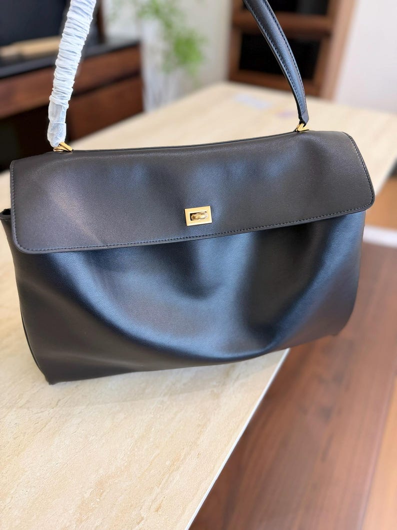 May include: Black leather handbag with a flap closure and a top handle. The bag features a gold-toned rectangular clasp on the front. The handle is attached with gold-toned hardware. The bag is sitting on a light-colored surface.