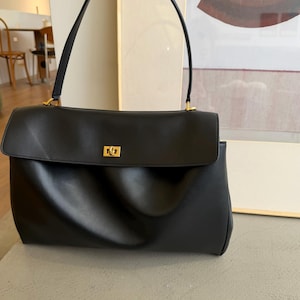 May include: Black leather handbag with a rectangular shape and a flap closure. The bag features a top handle and a gold-colored clasp. The design is minimalist and elegant, suitable for everyday use.