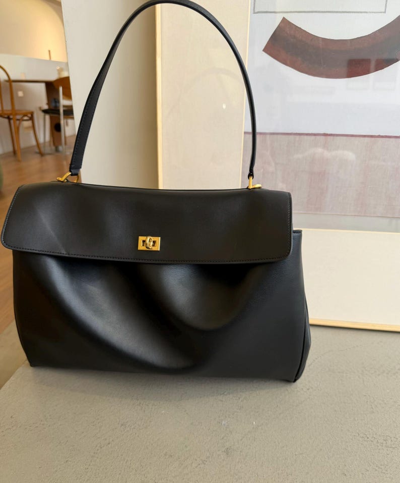 May include: Black leather handbag with a rectangular shape and a flap closure. The bag features a top handle and a gold-colored clasp. The design is minimalist and elegant, suitable for everyday use.