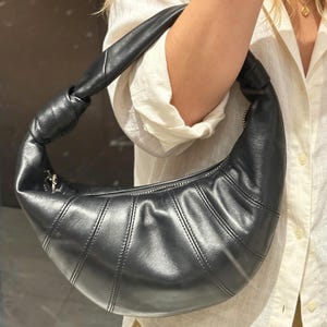 May include: A black crescent-shaped shoulder bag with a smooth, semi-glossy finish. The bag features a distinctive knotted handle, a curved silhouette, and vertical stitched panel detailing along the body, secured with a top zipper closure.