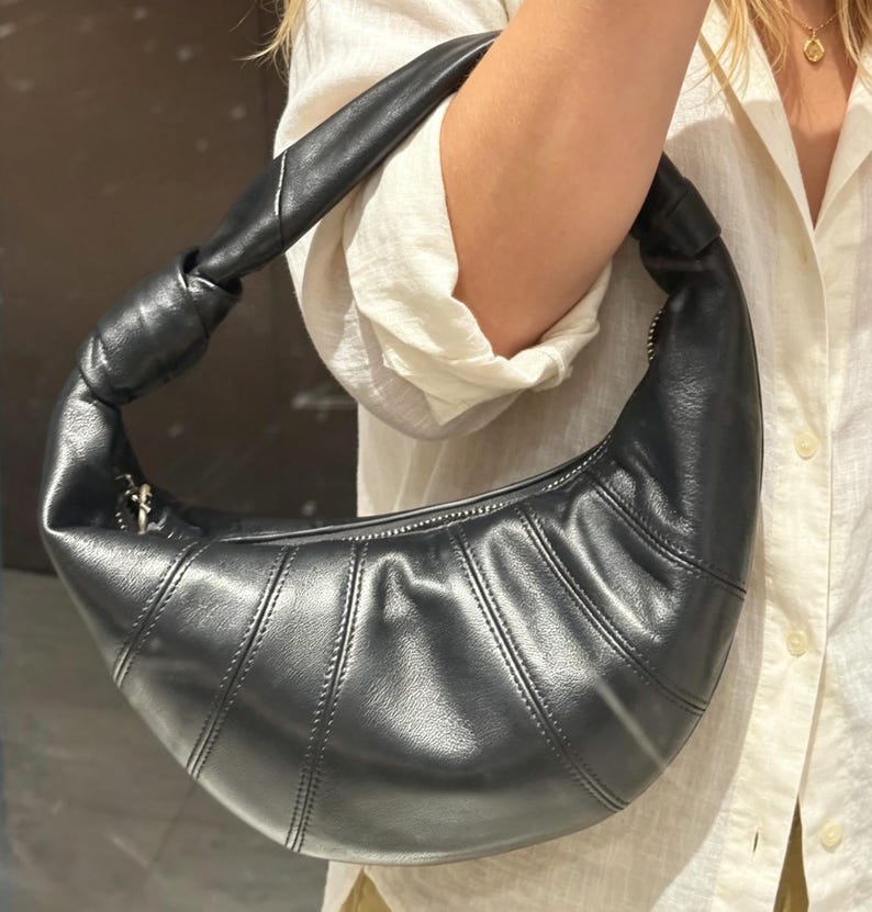 May include: A black crescent-shaped shoulder bag with a smooth, semi-glossy finish. The bag features a distinctive knotted handle, a curved silhouette, and vertical stitched panel detailing along the body, secured with a top zipper closure.