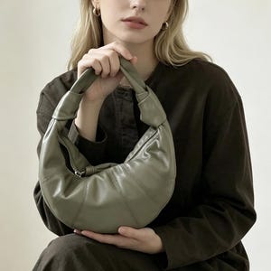 May include: A sage green crescent-shaped handbag with a smooth finish, featuring a knotted handle design and a silver-toned zipper closure along the top. The bag has a structured, paneled construction that curves into a rounded, slouchy silhouette.