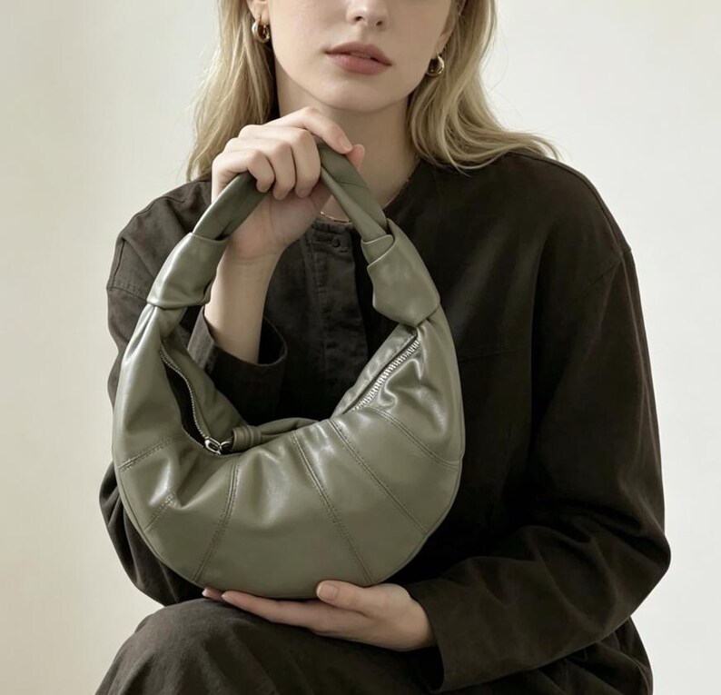 May include: A sage green crescent-shaped handbag with a smooth finish, featuring a knotted handle design and a silver-toned zipper closure along the top. The bag has a structured, paneled construction that curves into a rounded, slouchy silhouette.