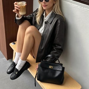 May include: A black leather jacket with a button-down front and a matching black handbag with a gold clasp. The image also shows black loafers, white socks, and sunglasses. A clear plastic cup with a beverage is held in the hand.