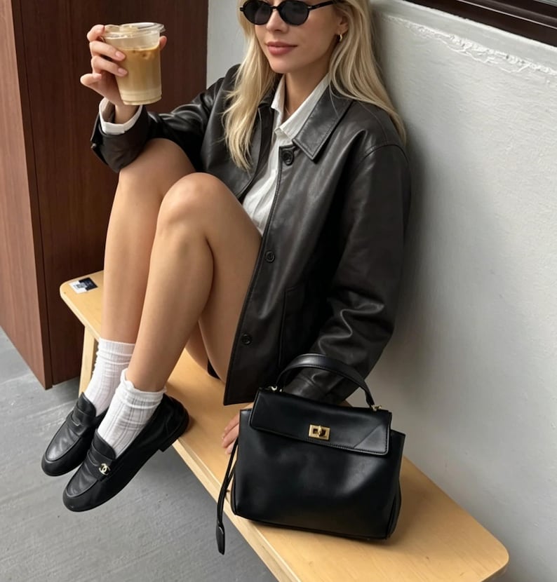 May include: A black leather jacket with a button-down front and a matching black handbag with a gold clasp. The image also shows black loafers, white socks, and sunglasses. A clear plastic cup with a beverage is held in the hand.
