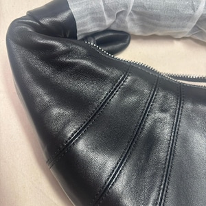 May include: A black leather shoulder bag with a curved, croissant-like silhouette. The bag features prominent vertical stitched seams, a silver-toned metal zipper closure, and a handle partially wrapped in protective white tissue paper.