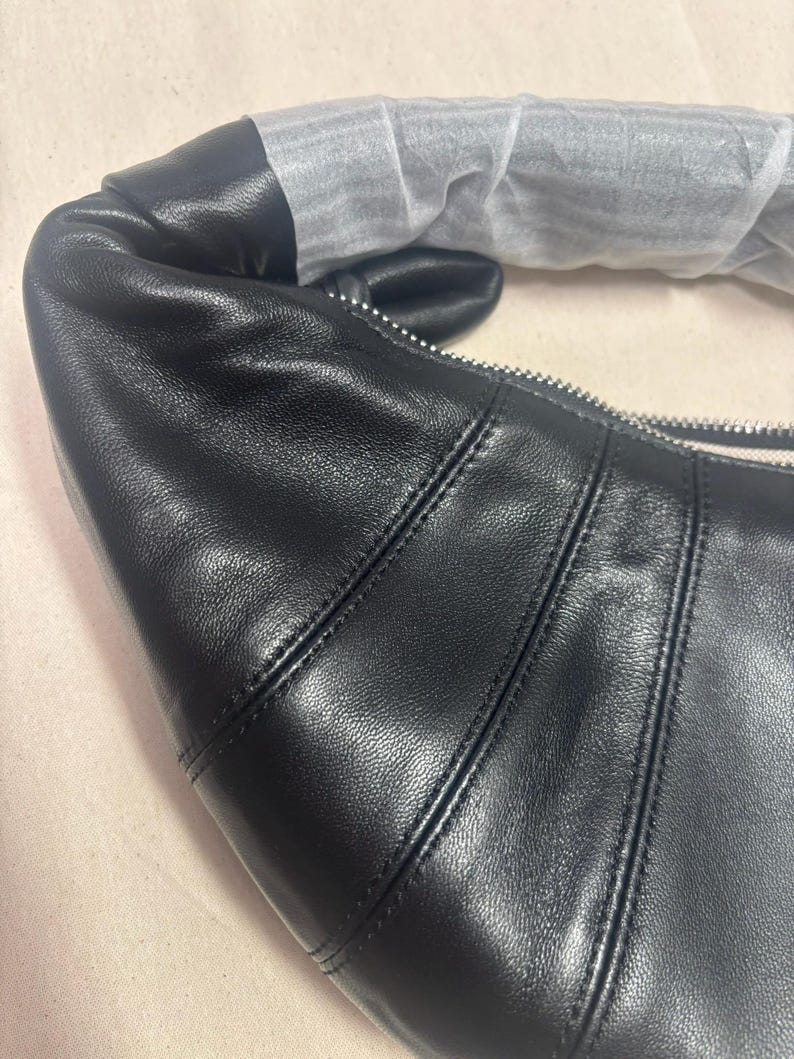 May include: A black leather shoulder bag with a curved, croissant-like silhouette. The bag features prominent vertical stitched seams, a silver-toned metal zipper closure, and a handle partially wrapped in protective white tissue paper.