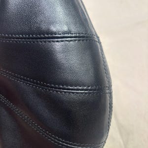 May include: A black leather handbag featuring a smooth, textured surface with prominent horizontal stitched seams. The material has a subtle sheen, and the construction displays clean, precise lines across the curved panel of the bag.