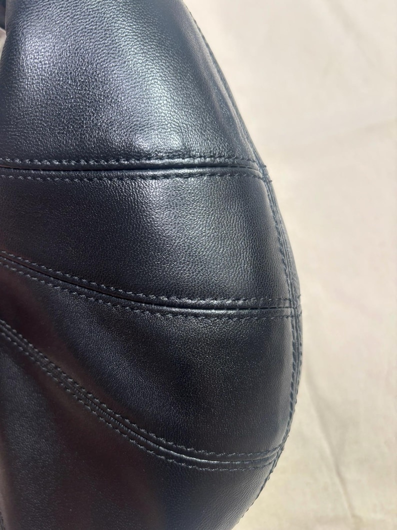 May include: A black leather handbag featuring a smooth, textured surface with prominent horizontal stitched seams. The material has a subtle sheen, and the construction displays clean, precise lines across the curved panel of the bag.