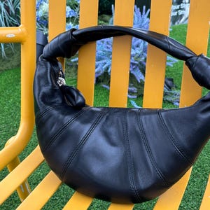 May include: A black crescent-shaped shoulder bag crafted from smooth leather with a distinctive knotted strap design. The bag features vertical stitched paneling across the body and a silver-toned metal zipper closure with a matching leather pull tab.