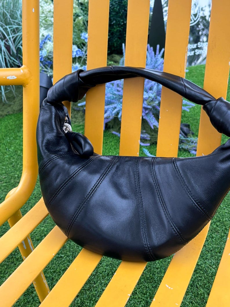 May include: A black crescent-shaped shoulder bag crafted from smooth leather with a distinctive knotted strap design. The bag features vertical stitched paneling across the body and a silver-toned metal zipper closure with a matching leather pull tab.