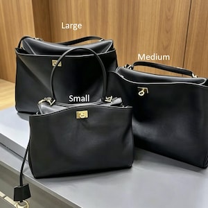 May include: Three black leather handbags of varying sizes: large, medium, and small. Each bag features a structured design with a top handle and a gold-colored clasp. The bags are displayed on a light gray surface.