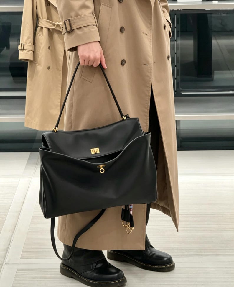 May include: A black leather handbag with a gold-colored clasp and handle attachments. The bag is held by a person wearing a tan trench coat and black boots. The bag has a rectangular shape with a flap closure.