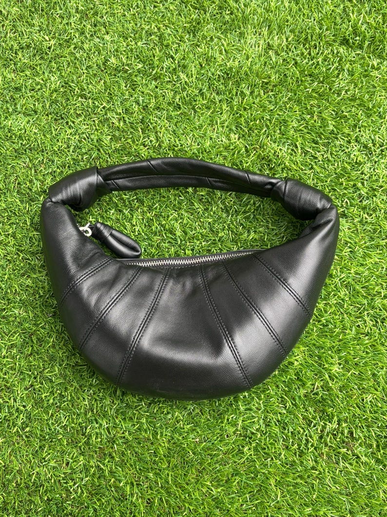 May include: A black crescent-shaped shoulder bag with a smooth finish, featuring a curved silhouette, a single integrated handle with knotted ends, and a silver-toned metal zipper closure along the top edge of the main compartment.