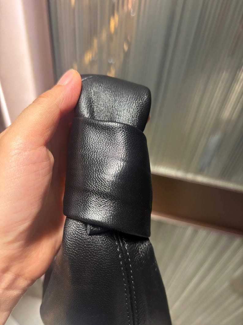 May include: A black handbag featuring a textured surface and a prominent knotted handle detail. The material has a visible grain and a matte finish, with clean stitching along the side seam. The design focuses on the structural knot at the top.