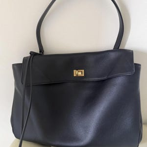 May include: Black leather tote bag with a rectangular shape and a top handle. The bag features a flap closure with a gold-colored metal clasp. A long, thin strap hangs from the top.
