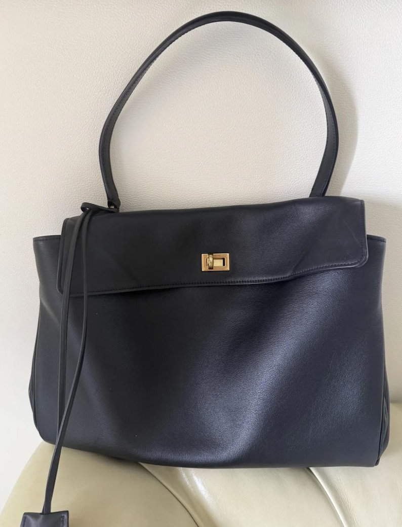 May include: Black leather tote bag with a rectangular shape and a top handle. The bag features a flap closure with a gold-colored metal clasp. A long, thin strap hangs from the top.