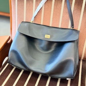 May include: A black leather handbag with a flap closure and a gold-toned rectangular clasp. The bag has a single shoulder strap and a white padded handle. The bag is sitting on a brown and white striped chair.