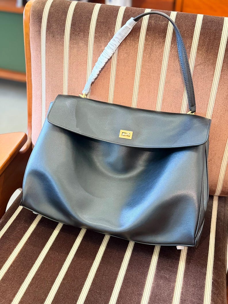May include: A black leather handbag with a flap closure and a gold-toned rectangular clasp. The bag has a single shoulder strap and a white padded handle. The bag is sitting on a brown and white striped chair.