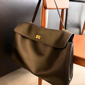 May include: A black leather handbag with a flap closure and a gold-tone clasp. The bag has a long shoulder strap and is hanging on a wooden chair. The bag is a classic style, suitable for everyday use.