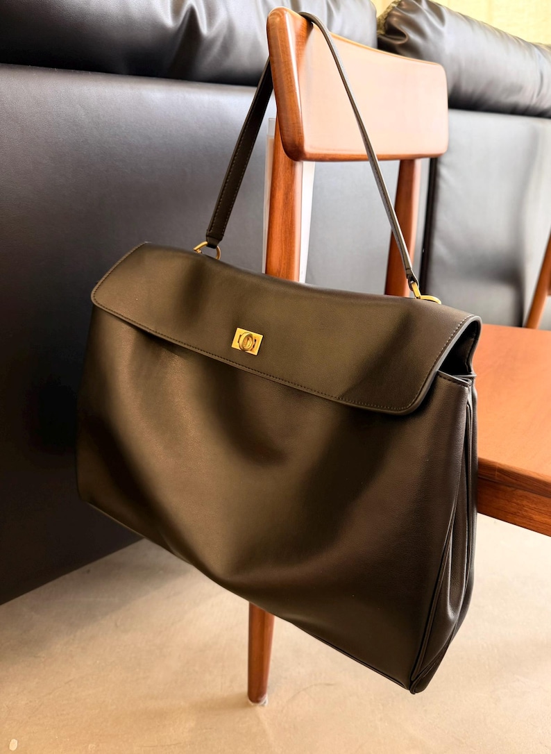 May include: A black leather handbag with a flap closure and a gold-tone clasp. The bag has a long shoulder strap and is hanging on a wooden chair. The bag is a classic style, suitable for everyday use.
