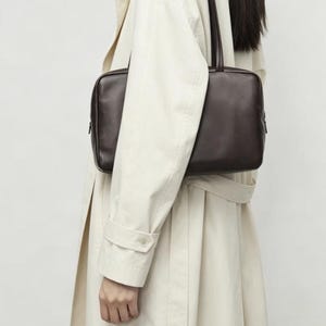 May include: A dark brown rectangular shoulder bag with a smooth finish, featuring a structured silhouette, rounded corners, and two thin, matching shoulder straps. The bag is designed with a clean, minimalist aesthetic and a secure top zipper closure.