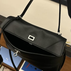 May include: Black leather shoulder bag with a rectangular flap and a silver-tone metal clasp. The bag has a long, thin strap and an open interior. The bag is shown with the flap open, revealing the interior.