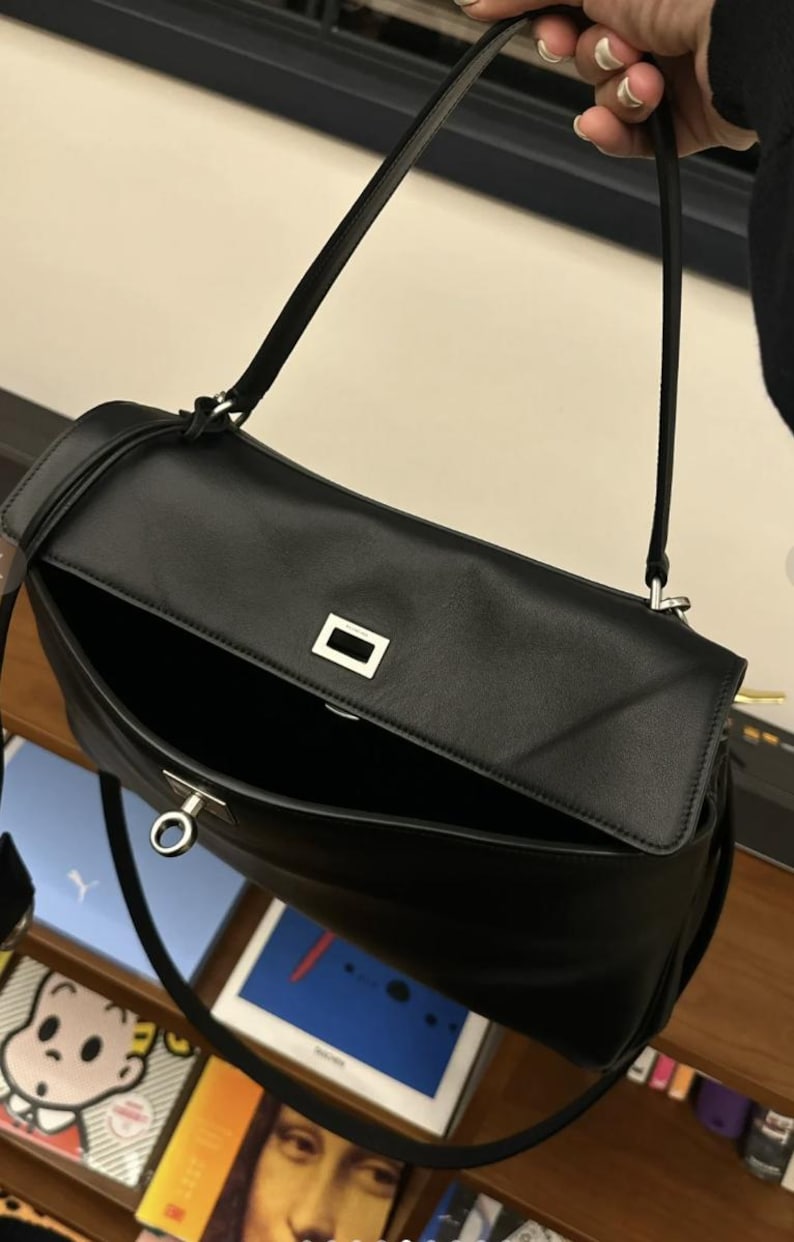 May include: Black leather shoulder bag with a rectangular flap and a silver-tone metal clasp. The bag has a long, thin strap and an open interior. The bag is shown with the flap open, revealing the interior.